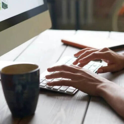 A picture showing hands typing on a keyboard to a computer screen. There's a black  coffee cup and a note book on the table too
